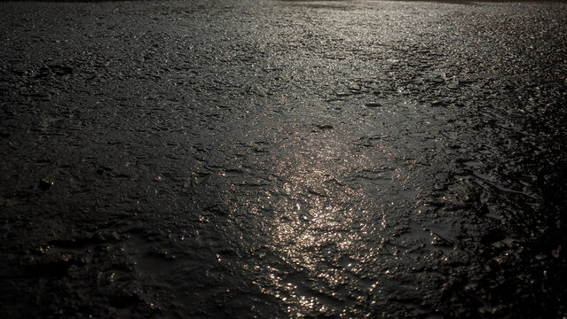 Wet dark rough asphalt pavement texture with bright sunlight reflection and small puddles.