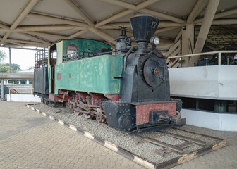 Historic steam locomotive displayed under a covered railway structure, showing its weathered metal body, wheels, and mechanical components. © Nuzul