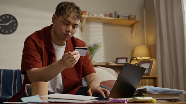 Asian young adult man holding credit card and typing on laptop at home, paying utility bills. Footage is suitable for marketing campaigns of online banking, e commerce, and fintech services