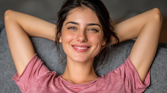 A smiling woman with dark hair and blue eyes relaxes on a gray sofa with her hands behind her head against a blurred background.