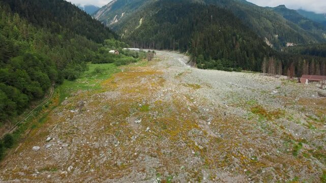 Dramatic aerial photography flying over the remnants of a devastating landslide in Georgia's remote Shovi Racha mountain valley, revealing a wide path of rocks and debris among lush green coniferous f
