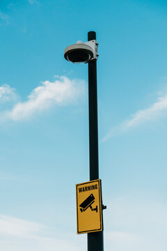 A surveillance camera and warning sign stand on a pole against a blue sky. Security, monitoring, privacy and public safety concept.