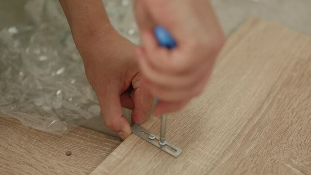 Hands assembling furniture at home, using a screwdriver to secure metal brackets to wooden panels on a light-colored surface with plastic wrapping visible