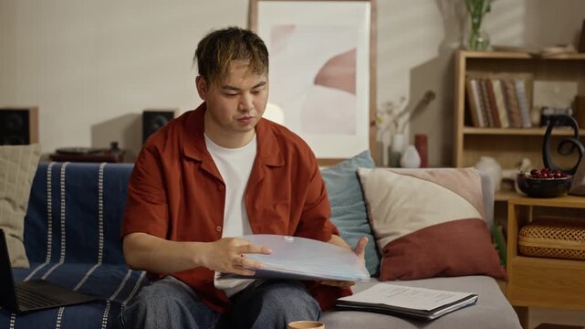 Young adult Asian man sitting on sofa, organizing utility bills and financial documents from expanding folder. Footage is perfect for marketing personal finance apps or banking services