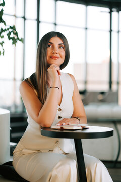 Young woman in a white vest sitting at a cafe table thinking during the day in a bright room