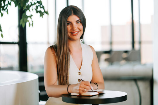 Professional woman with long dark hair smiling while writing in a notebook at a bright indoor office