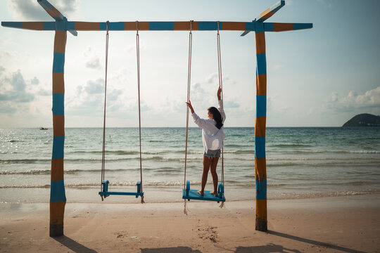Fit Asian woman stands barefoot on a beach swing, experiencing relaxation and self-care in paradise