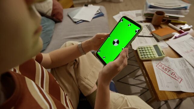 Young adult Caucasian woman holding smartphone with green screen and tracking markers near table with past due bills. Video is ideal for promoting mobile banking, debt relief, or budgeting apps