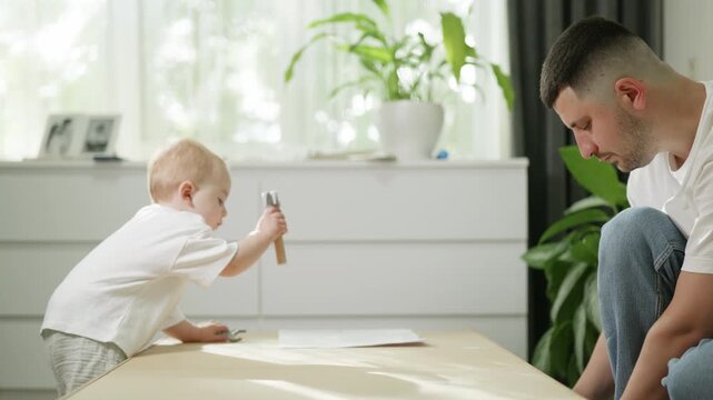 Father and toddler assembling furniture at home, child holding hammer while parent focuses on wooden panel, bright interior with plants and drawers in the background