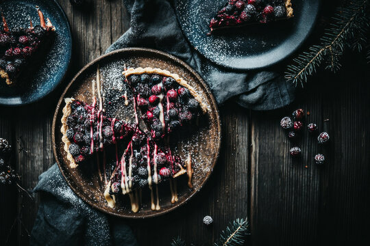 Blueberry pie is set on a wooden plate. The pie has pieces cut out and shows blueberries and a vanilla drizzle. It is placed on a dark surface, which enhances its colors