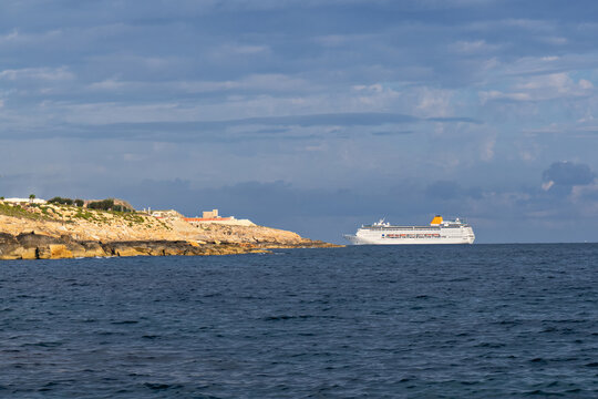 White cruise ship sailing beside rocky coastline across dark blue sea, dramatic layered clouds creating scenic maritime travel atmosphere