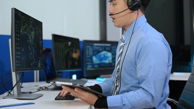 Asian male dispatcher with headset uses a tablet and points at flight radar screens