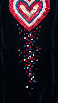 A heart shape made of red, white, and blue sequins on a black background, symbolizing patriotism and Valentine's Day, with scattered sequins below.