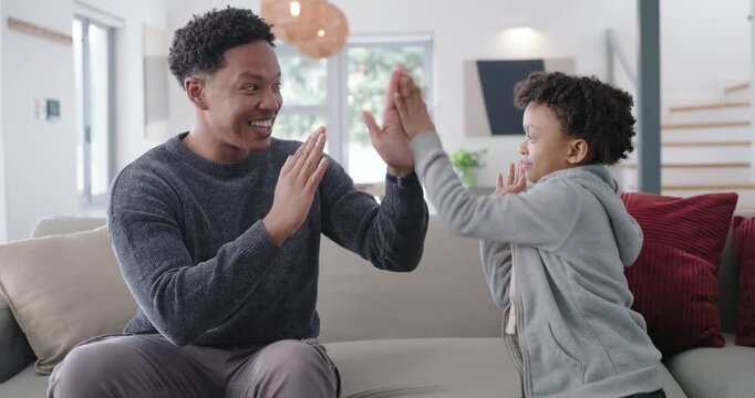 African American man initiating hand-clap game, child mirroring on living room sofa for bonding