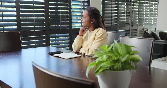African American woman sitting by shutters at home, gazing then writing notebook with pen, planning