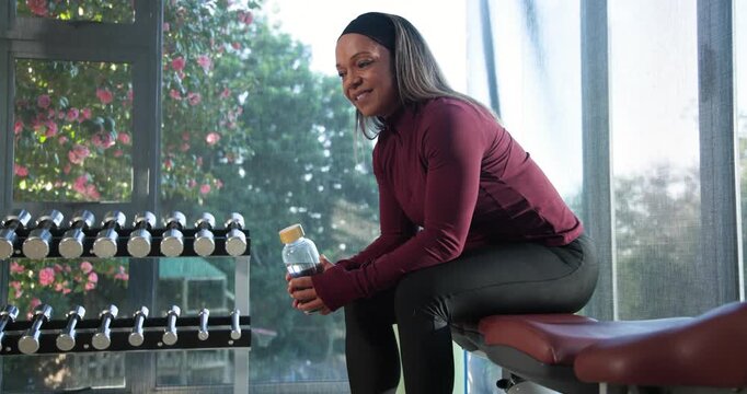 Mature African American woman sitting on bench holding water bottle, rehydrating after set, smiling