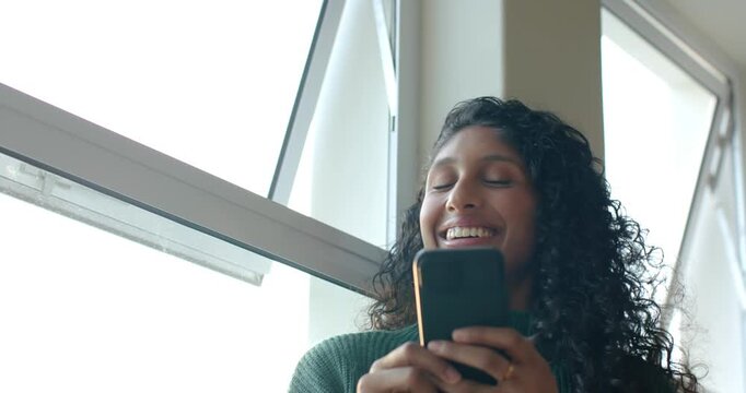 African American woman sitting by slanted window holding smartphone, scrolling and laughing softly
