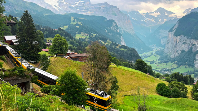 Yellow Mountain Train Above a Lush Swiss Alpine Valley. Yellow mountain train travels through green Swiss alpine meadows above a dramatic valley with steep cliffs, forests, chalets and snowcapped