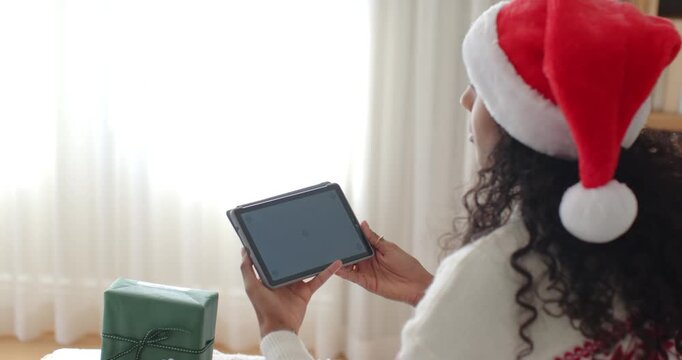 African American woman holding tablet near window in Santa hat, tilting to avoid glare beside gift