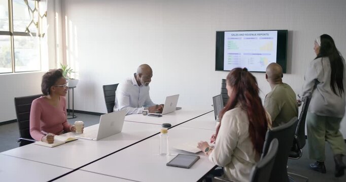 Diverse team sitting at table watching presenter in blazer walking in, urging focus on sales charts