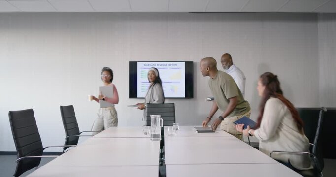 Diverse team entering from right, placing laptops, pitcher at table, preparing Sales slide