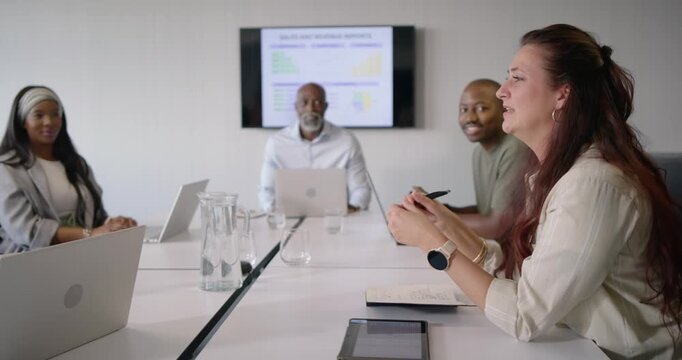Diverse coworkers responding to sales slide at table, redhead woman leaning, gesturing with pen