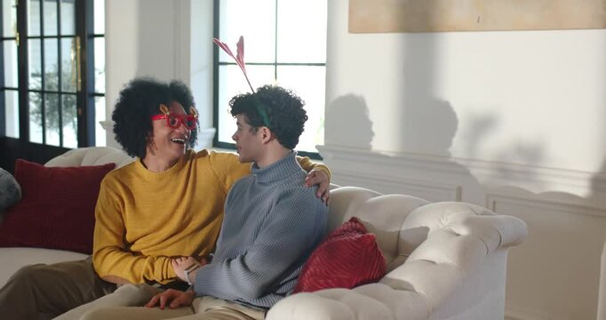 African American men laughing holding hands leaning on sofa in living-room sparked by heart glasses