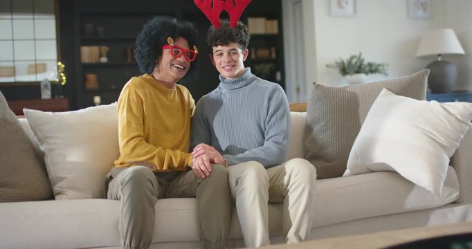 Holding hands, two men on sofa at home adding red glasses and antler headband, posing portrait