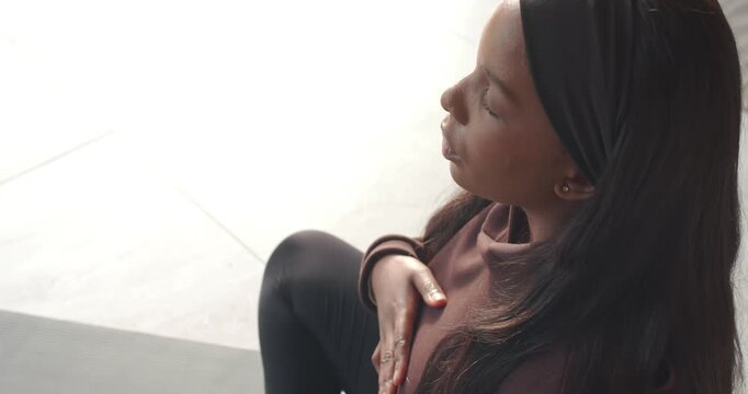 African American woman bringing hand to chest, practicing breathwork on mat in bright room