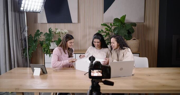 Diverse female cohosts looking at camera and white box, lifting lid and unboxing items for viewers