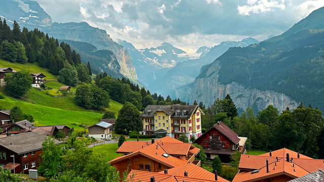 Swiss Alpine Village Overlooking a Dramatic Mountain Valley. Swiss alpine village with wooden chalets and red roofs overlooking a lush mountain valley beneath dramatic cloudy peaks.