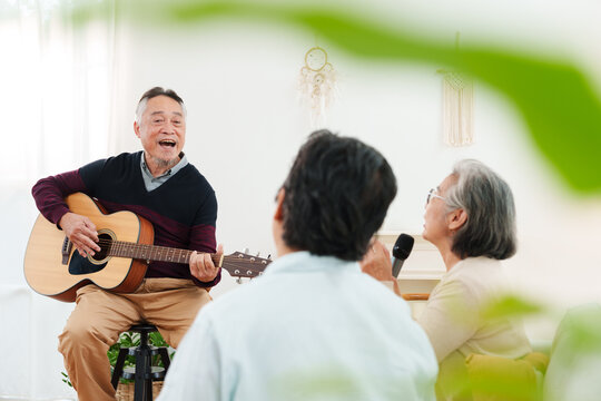 Cheerful, joyful Asian senior friends spending leisure time together. Elderly man playing acoustic guitar while senior women sitting on sofa singing along in white cozy, modern living room at home