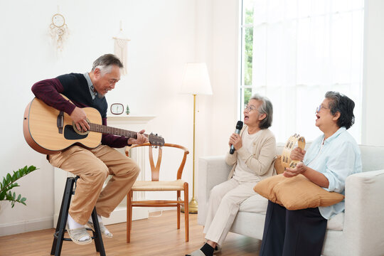 Cheerful, joyful Asian senior friends spending leisure time together. Elderly man playing acoustic guitar while senior women sitting on sofa singing along in white cozy, modern living room at home
