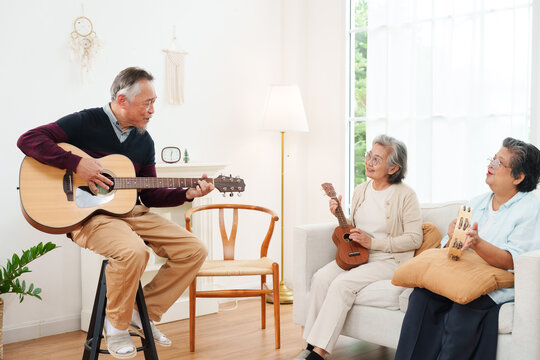 Cheerful, joyful Asian senior friends spending leisure time together. Elderly man playing acoustic guitar while senior women sitting on sofa singing along in white cozy, modern living room at home
