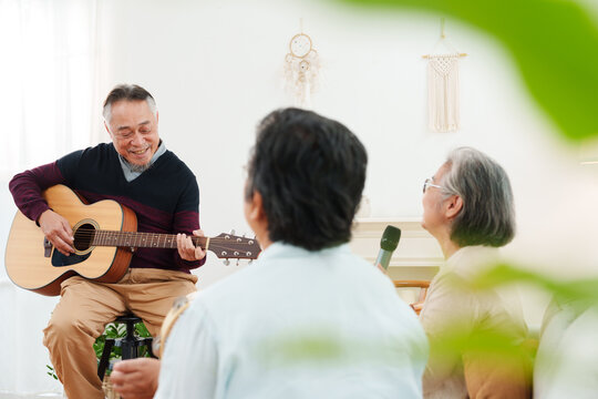 Cheerful, joyful Asian senior friends spending leisure time together. Elderly man playing acoustic guitar while senior women sitting on sofa singing along in white cozy, modern living room at home