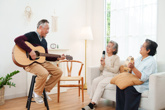 Cheerful, joyful Asian senior friends spending leisure time together. Elderly man playing acoustic guitar while senior women sitting on sofa singing along in white cozy, modern living room at home