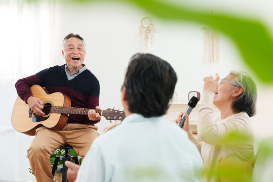 Cheerful, joyful Asian senior friends spending leisure time together. Elderly man playing acoustic guitar while senior women sitting on sofa singing along in white cozy, modern living room at home