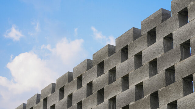 Diagonal view of a modern grey concrete cinder block wall with repetitive hollow pattern set against a blue sky with soft white clouds.