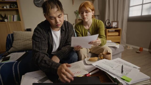 Young adult Asian man opening laptop as camera tilts up from past due utility bills with Caucasian woman nearby. Perfect for debt relief and financial planning campaigns