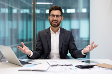 Fototapeta premium Portrait of a young confident businessman in a suit sitting in the office at a desk in front of the camera, smiling and spreading his arms, inviting and introducing