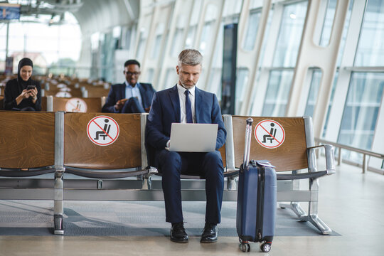 A man in a suit sits in an airport waiting area, focused on his laptop, with social distancing markers on adjacent seats