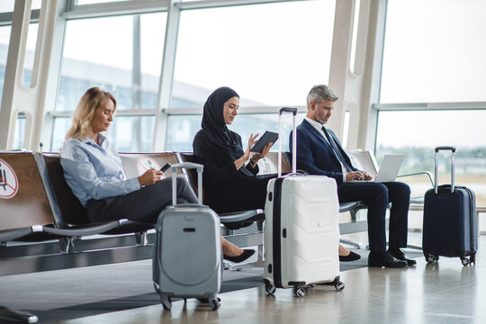 People of different ethnicities and genders sit in an airport terminal, engaged with their phones, tablets, and laptops while their suitcases are nearby