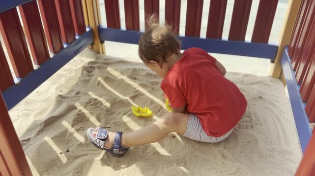 Toddler sitting in a sandbox and playing with a red bucket at a playground, with sand and wooden rails around the play area