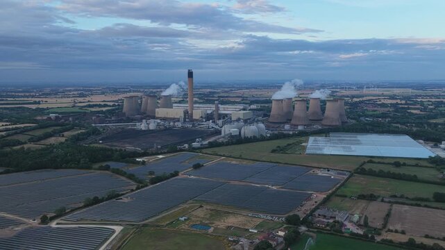 Large industrial power station with cooling towers standing near rows of solar panels in the countryside during the golden hour as smoke drifts slowly from the tall chimney into the evening sky.