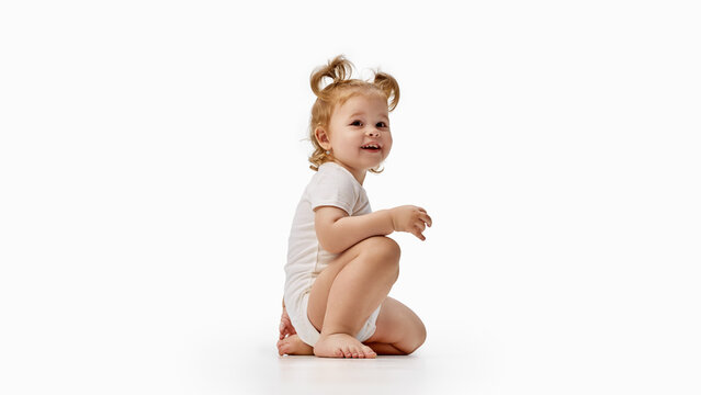 Smiling toddler girl kneeling in white bodysuit. Concept of happiness, flexible movement, posture and early childhood development, for parenting and childcare.