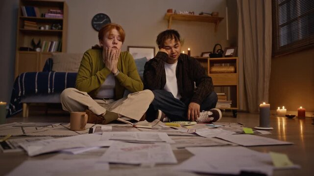 Young adult Caucasian woman and Asian man sitting on floor surrounded by utility bills, looking stressed and overwhelmed by financial debt. Clip is ideal for financial planning or debt relief use