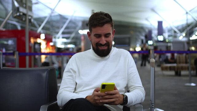 Young caucasian man with luggage using his phone sending online messages while waiting for a flight at the airport gate. Video with copy space.