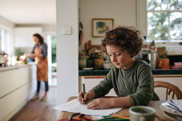 Fototapeta premium A young child intently draws on paper in a cozy kitchen while a parent prepares food in the background, capturing a moment of creativity and domestic life.