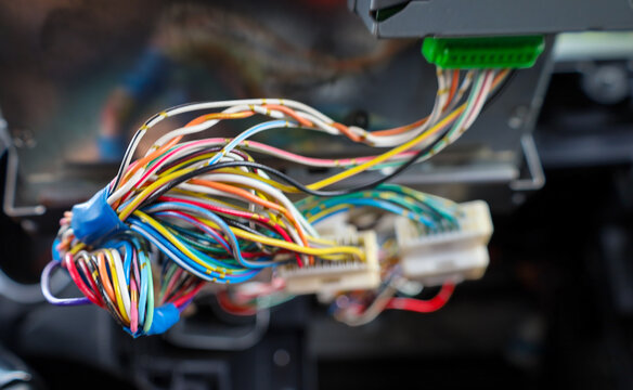 ​A close-up bundle of colorful electrical wires with plastic connectors hangs from a disassembled car dashboard. The auto electronics repair process shows complex cable wiring during onboard system ma