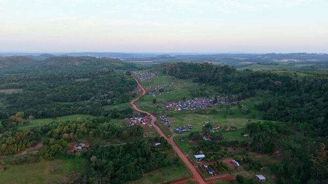 Drone footage shows a large crowd gathered along a rural dirt road for WRC Rally 2026 in Nueva Alborada, Paraguay. Colorful campsites and scenic forested hills,